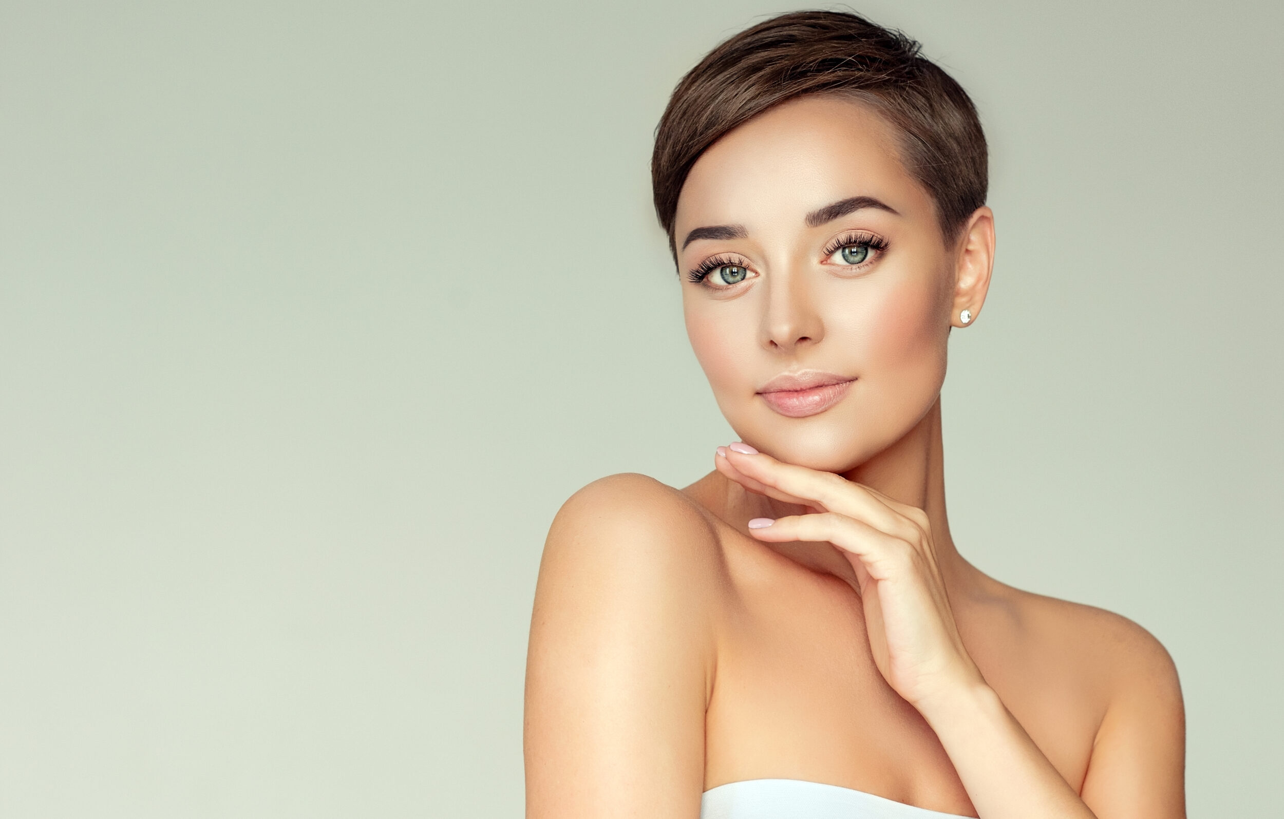 Young, brown haired woman with short stylish haircut, clean fresh skin and light, delicate makeup is touching the face by slender fingers. Cosmetology.